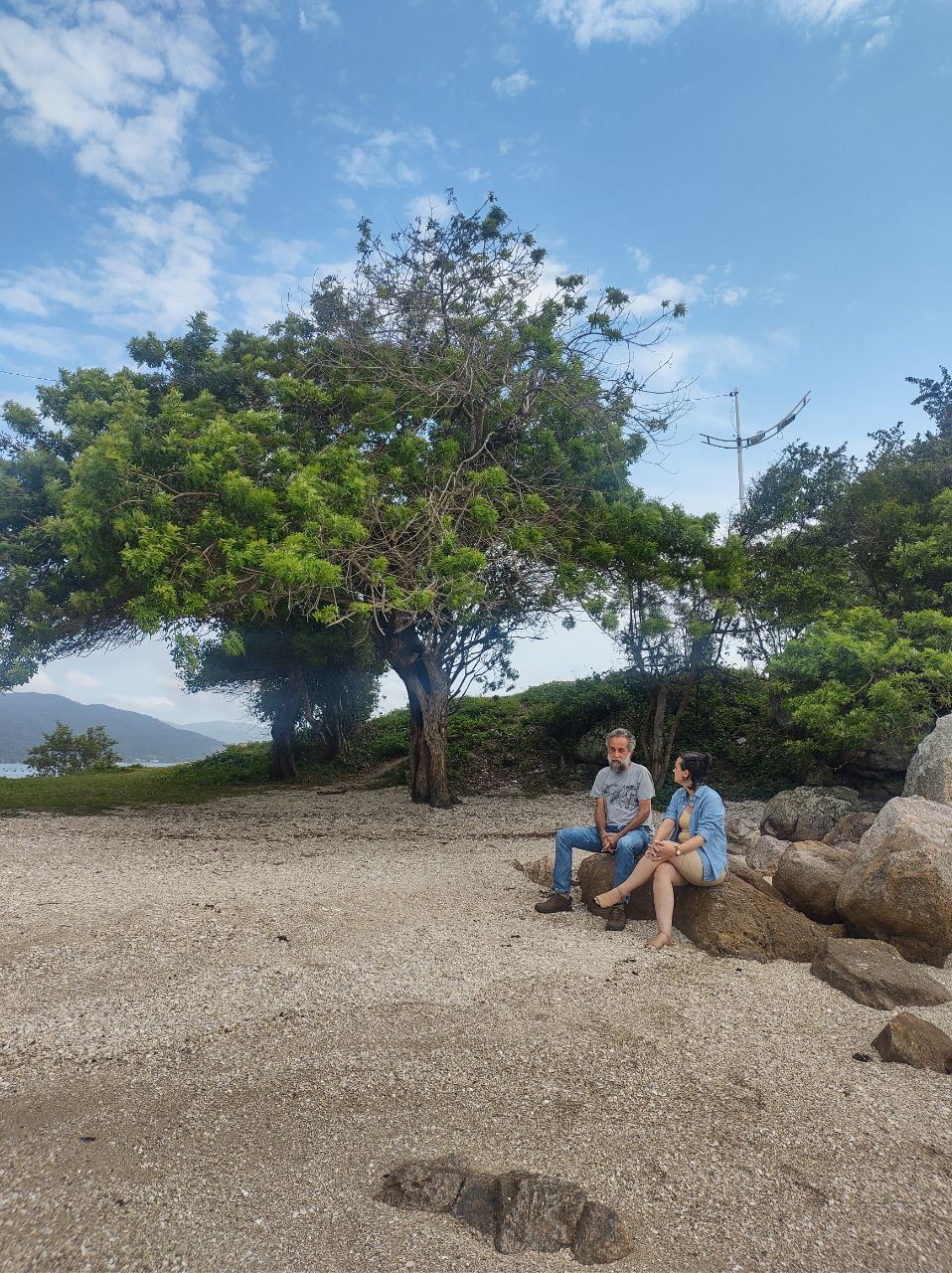 Beatriz Costa e o arqueólogo Lucas Bueno conversam sentados nas pedras de uma praia em Florianópolis