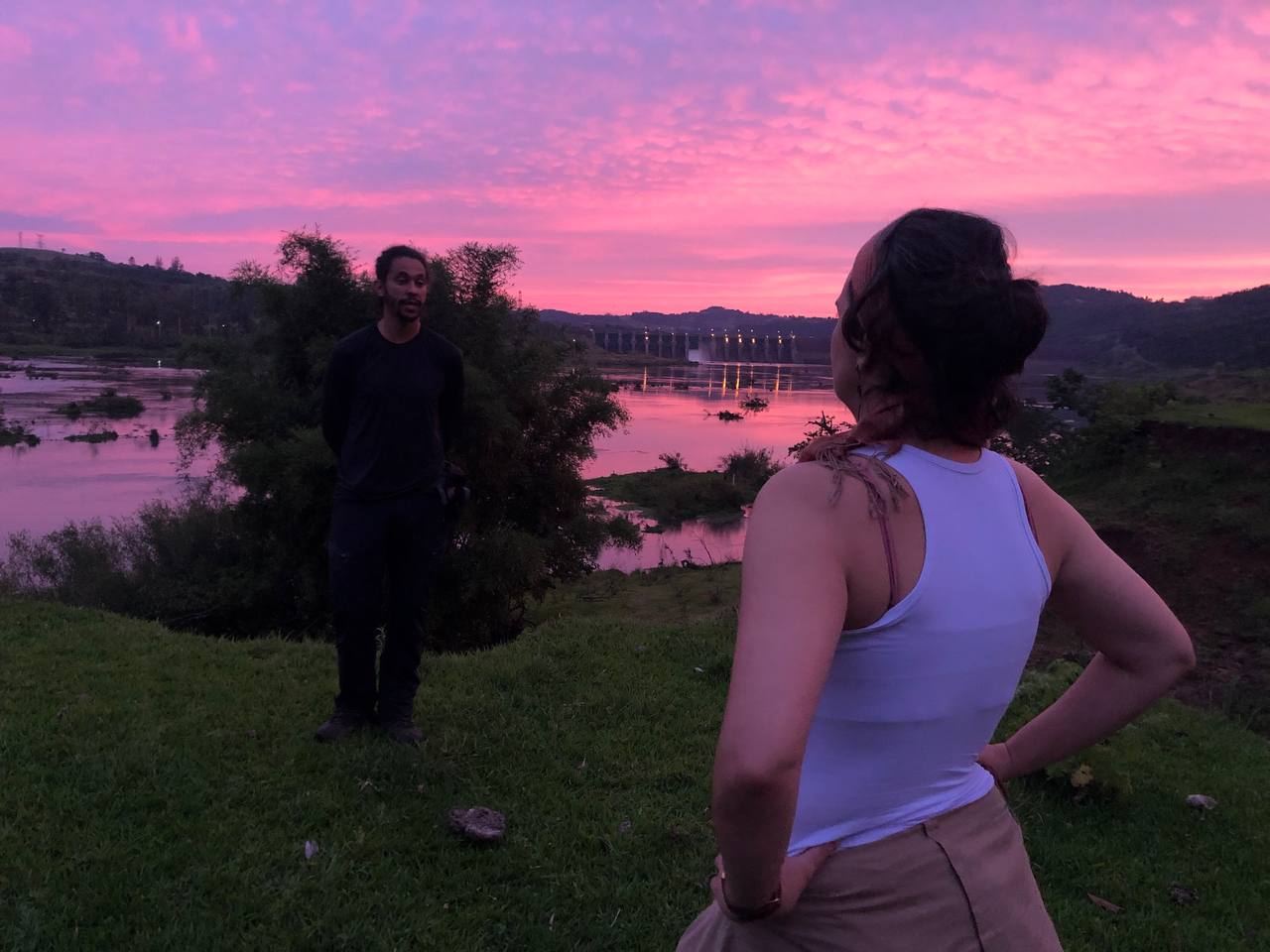Gravação ao entardecer, com céu rosa, à beira de uma represa: Beatriz Costa conversa com o arqueólogo Marcos Santos